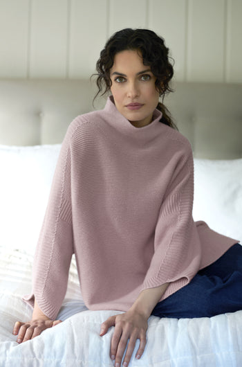 A person with dark curly hair sits on a bed wearing a light pink Merino Wool Stitch Detail Jumper and blue pants, gazing directly at the camera.