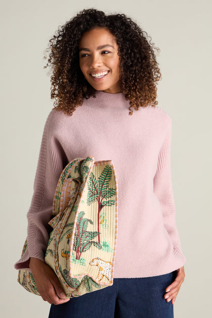 A woman with curly hair smiles while holding a patterned bag. She is wearing a light pink Merino Wool Stitch Detail Jumper and denim pants, exuding cozy comfort.