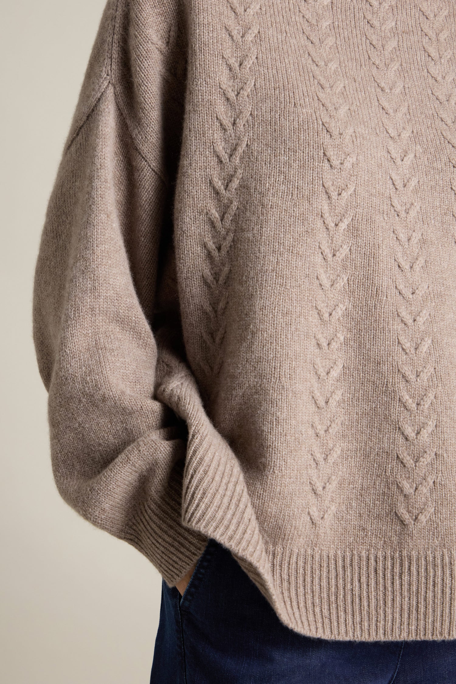 A person wearing a beige Cashmere Blend Cable Jumper with their hand in the pocket of dark blue jeans.