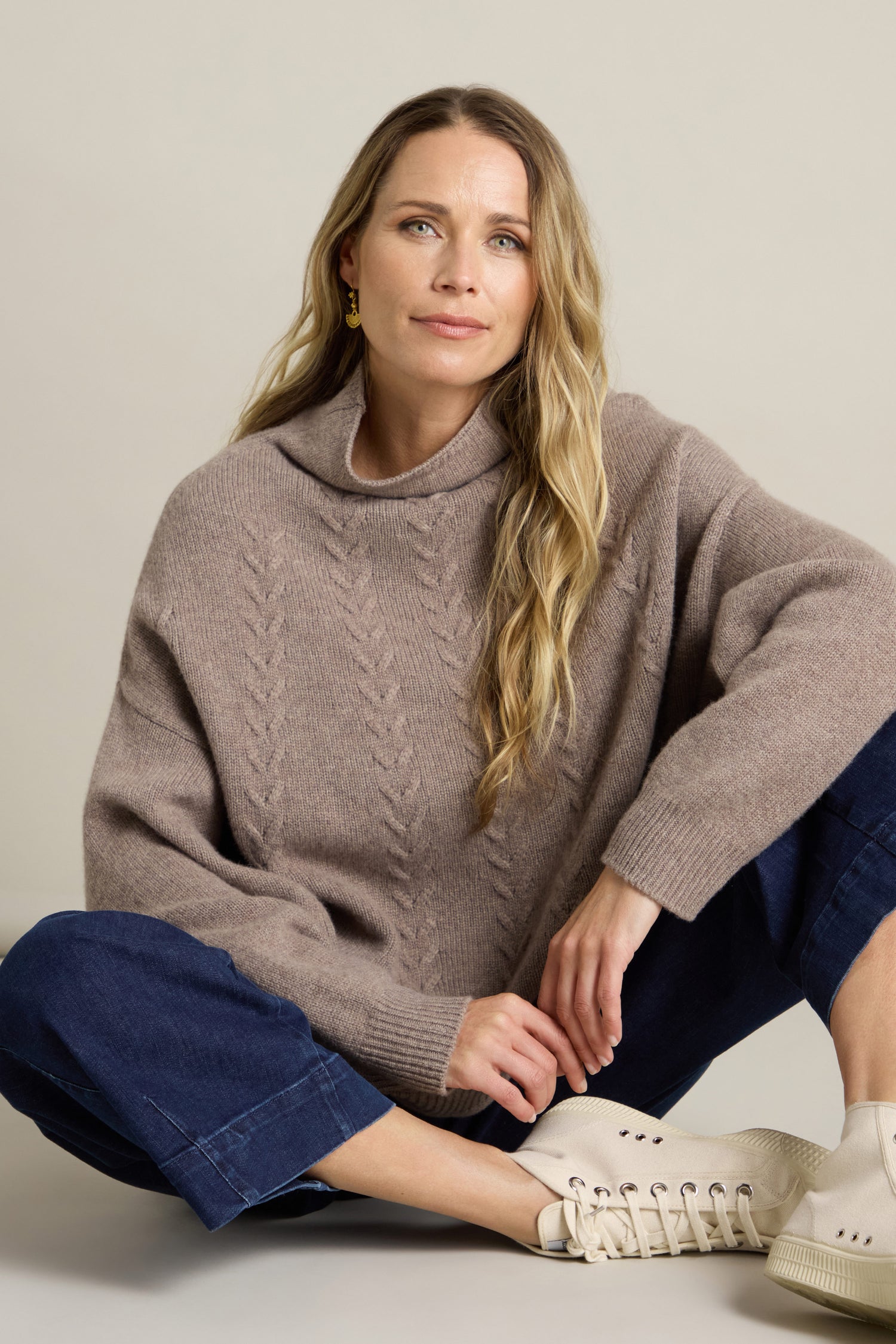 A woman with long blonde hair wears a light brown Cashmere Blend Cable Jumper, blue jeans, and white sneakers. She sits on the floor with one knee bent, resting her hand on it, and gazes slightly upwards.