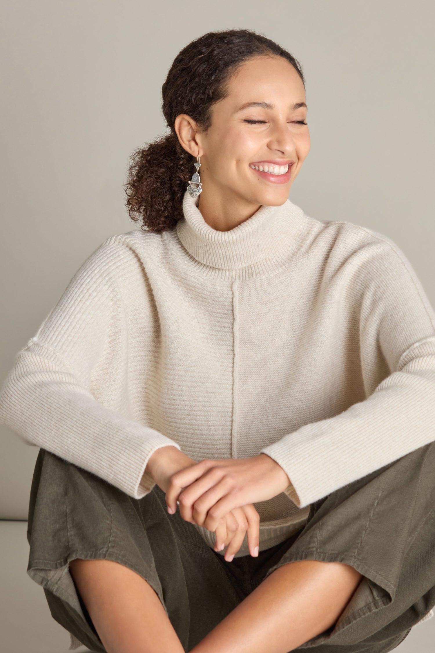 A person wearing a cream-colored Merino Wool Boxy Jumper and olive-green pants is sitting cross-legged, smiling, and looking to the side.