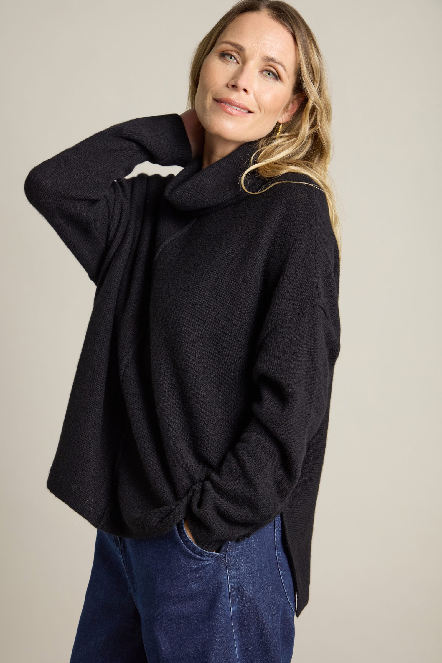 A person stands against a plain background, wearing a black Merino Wool Cowl Neck Jumper and blue jeans. They have one hand in their pocket and the other resting near their neck.