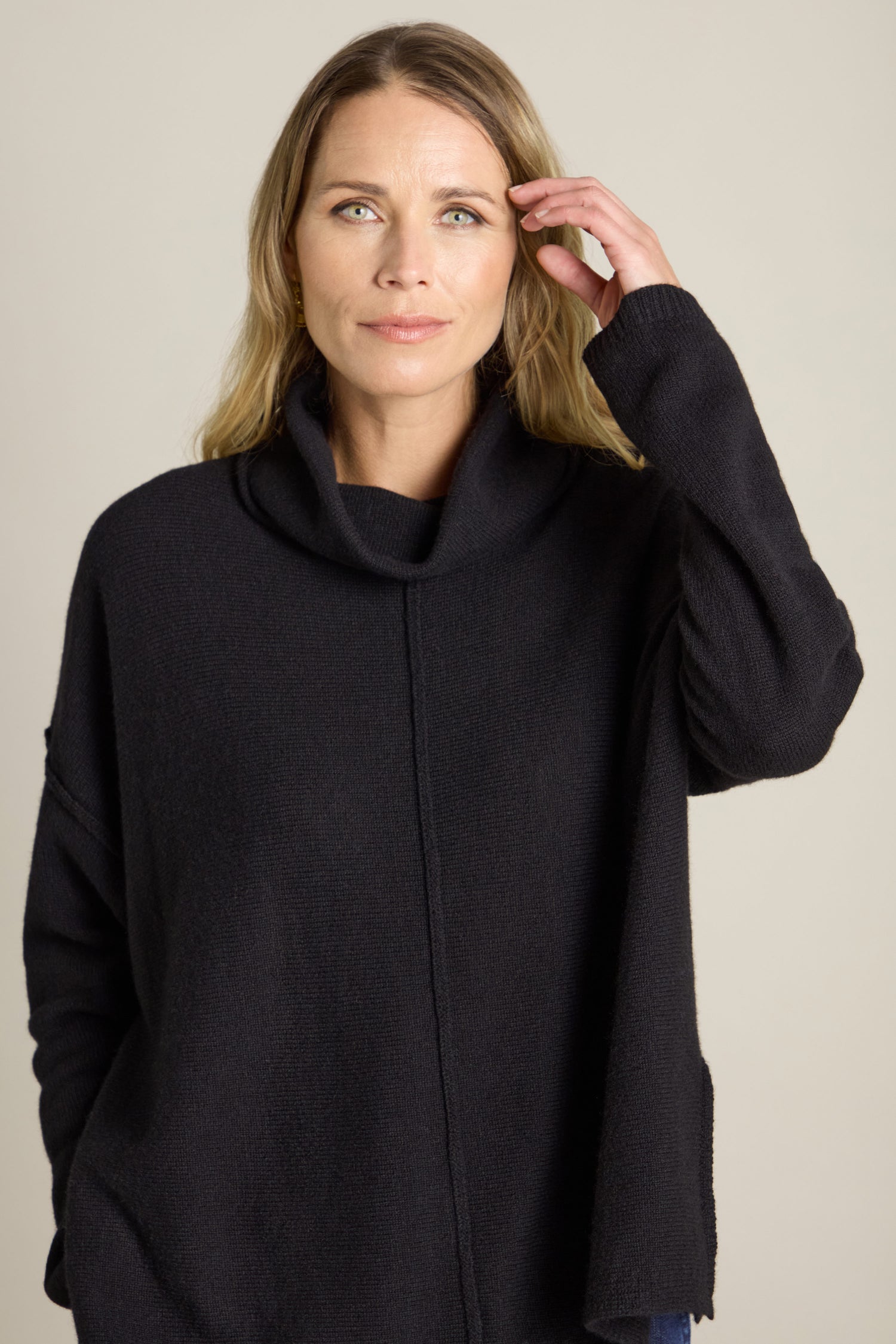 A woman with long hair is wearing a loose-fitting, extra fine Merino Wool Cowl Neck Jumper in black. She is looking directly at the camera and touching her hair with her right hand. The background is plain and neutral.