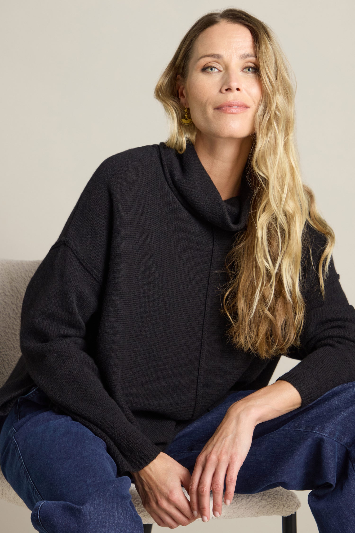 A woman with long blond hair wearing a Merino Wool Cowl Neck Jumper and blue pants sits on a beige chair against a plain background.