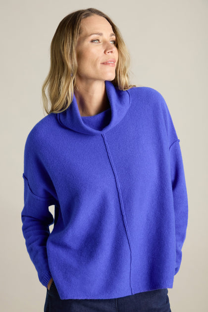 A person with long, wavy blonde hair wears a bright blue Merino Wool Cowl Neck Jumper, looking to the side.