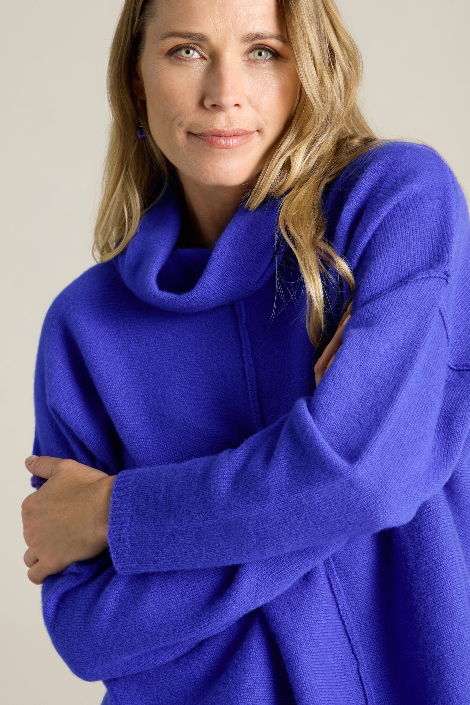 A woman with long blonde hair wearing a Merino Wool Cowl Neck Jumper stands with her arms crossed, looking at the camera.