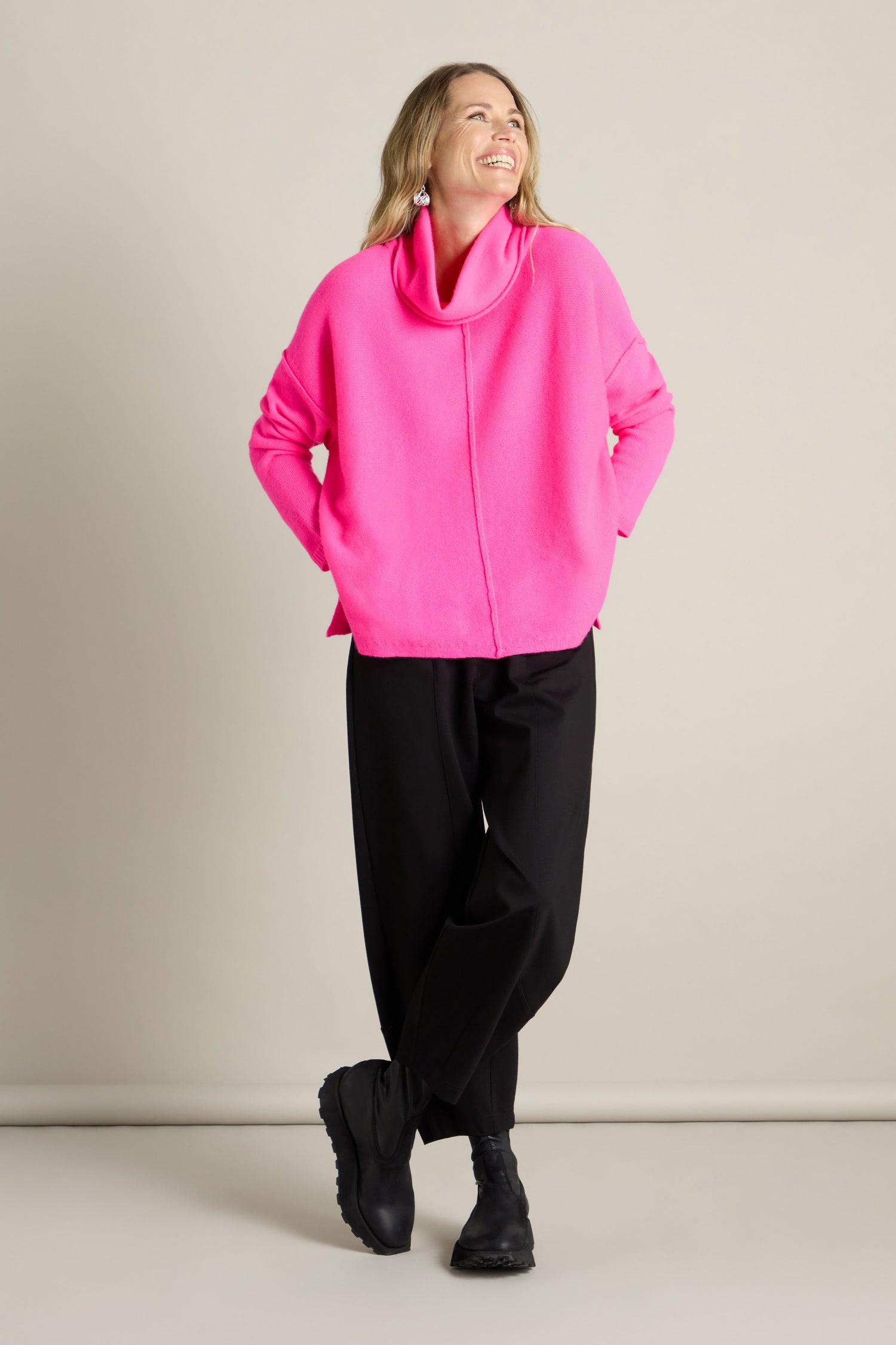 A person standing against a plain background, wearing a bright pink Merino Wool Cowl Neck Jumper, black pants, and black shoes, with a relaxed, smiling expression.