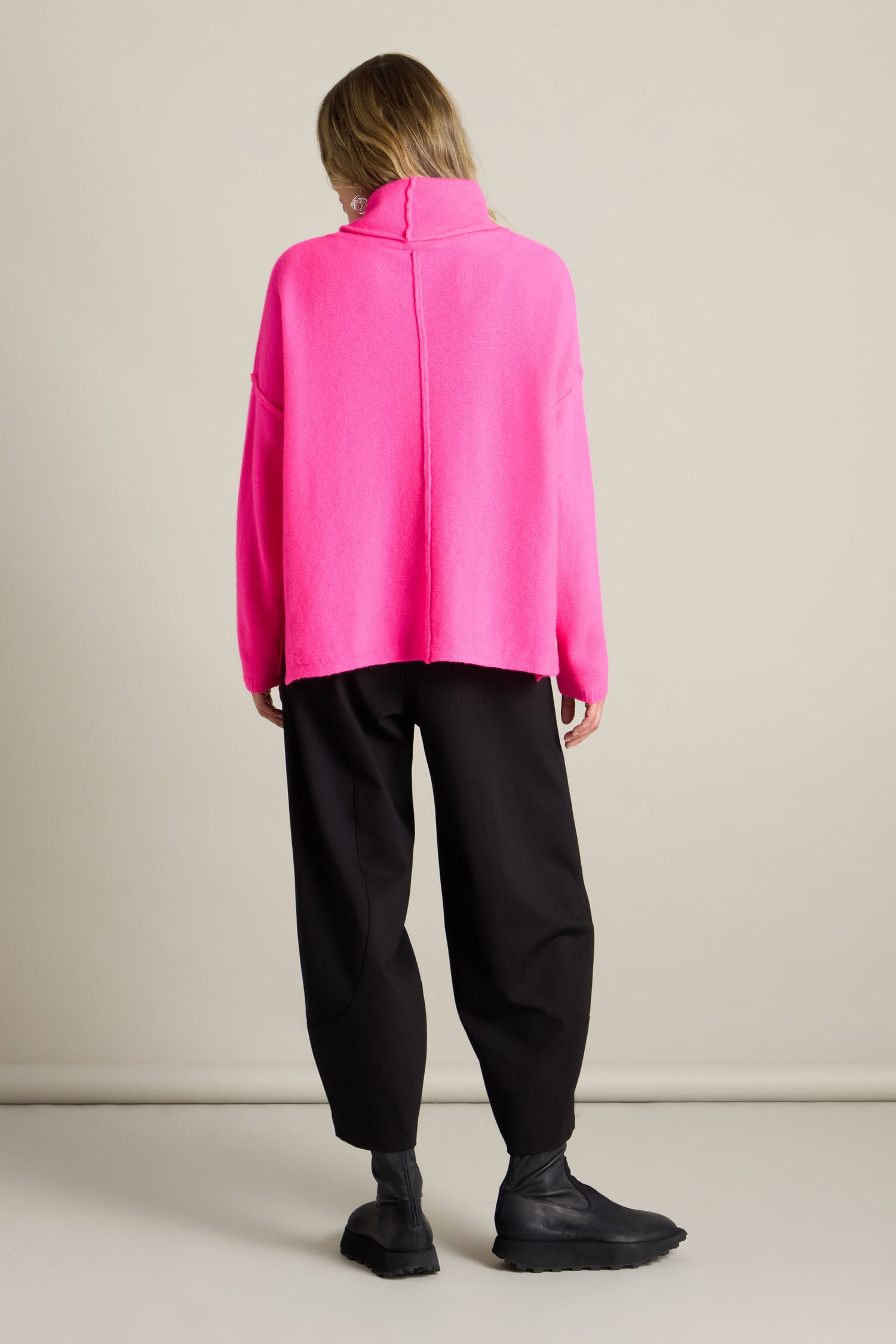 Person with long hair wearing a bright pink Merino Wool Cowl Neck Jumper, black baggy pants, and black boots, standing facing away from the camera against a plain background.