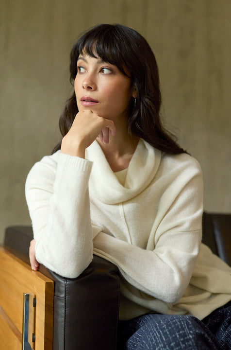 A woman with long dark hair and bangs sits on a dark leather sofa, resting her chin on her hand and gazing thoughtfully to the side. She is wearing a women’s Merino Cowl Neck Jumper in white and dark pants.
