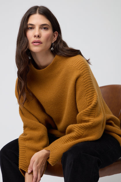 A woman with long dark hair wears the Merino Wool Stitch Detail Jumper in mustard, featuring a relaxed boxy silhouette. She stands against a plain light gray background, facing the camera.