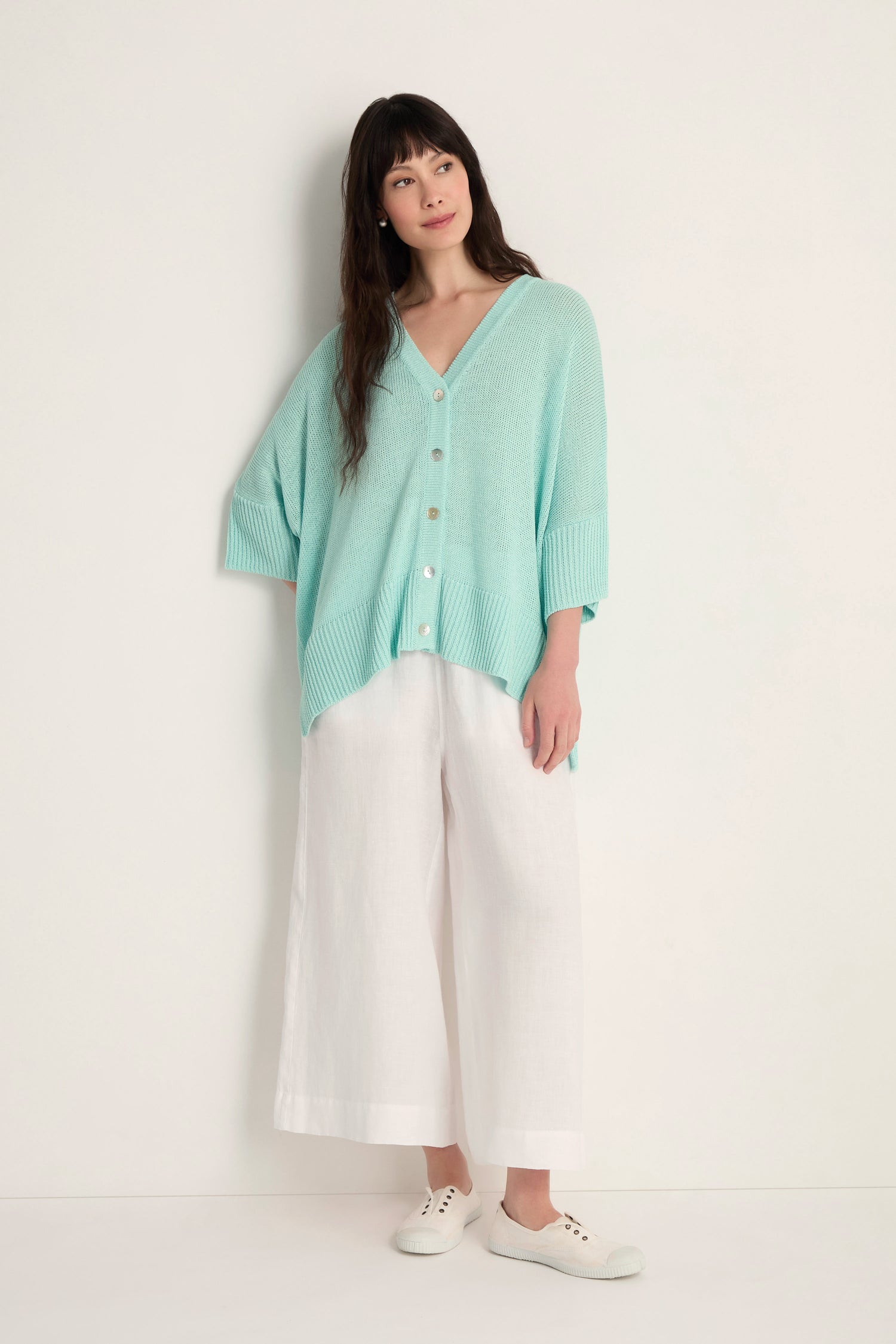 A woman stands against a white wall wearing the Relaxed Aqua Cardigan with buttons, paired with white wide-leg pants and sneakers for an effortless and lightweight layered look.