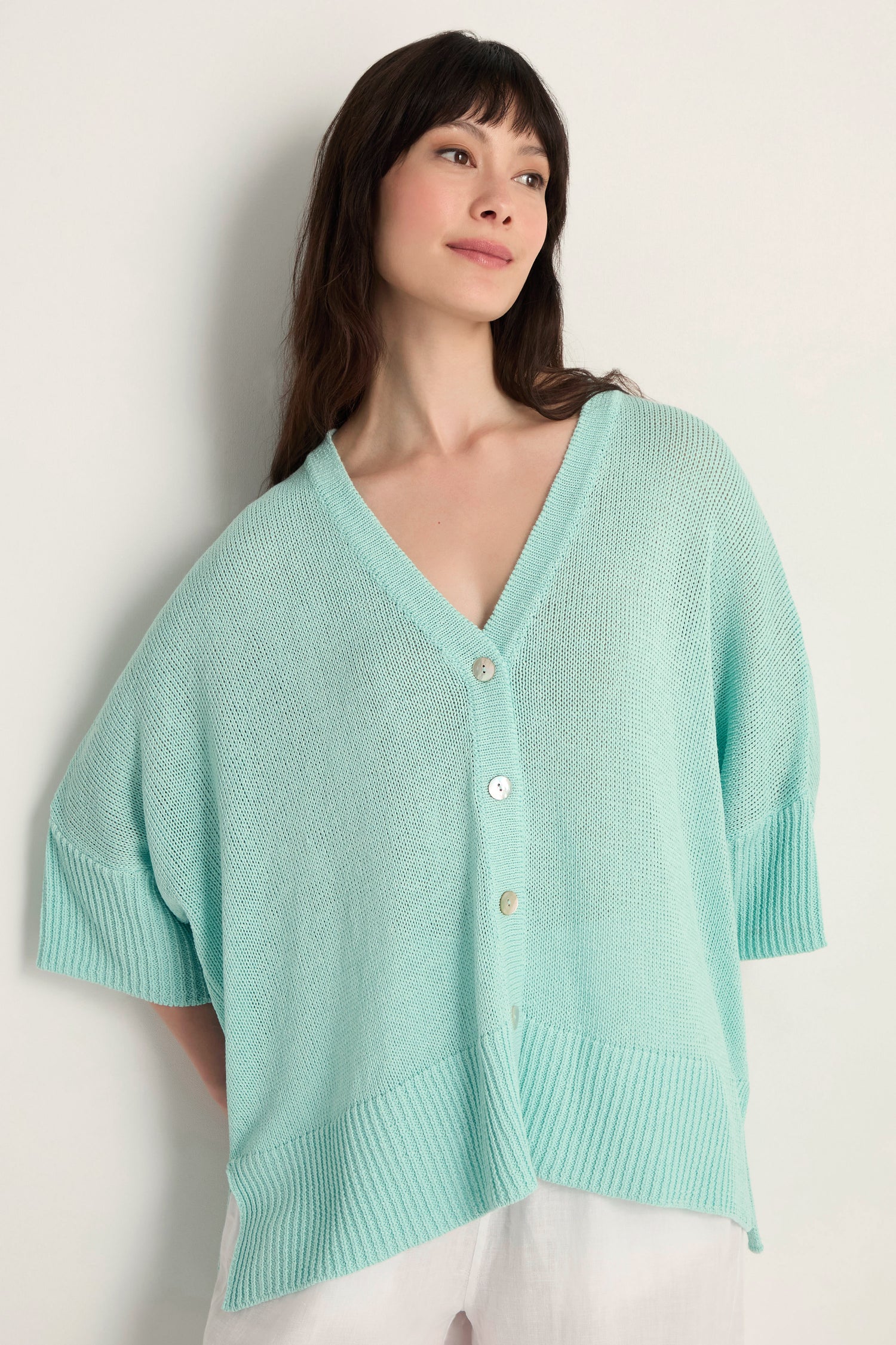 A woman wears the Relaxed Aqua Cardigan, a light blue open knit design with white buttons, layered over a white outfit—ideal for lightweight layering against a plain light background.