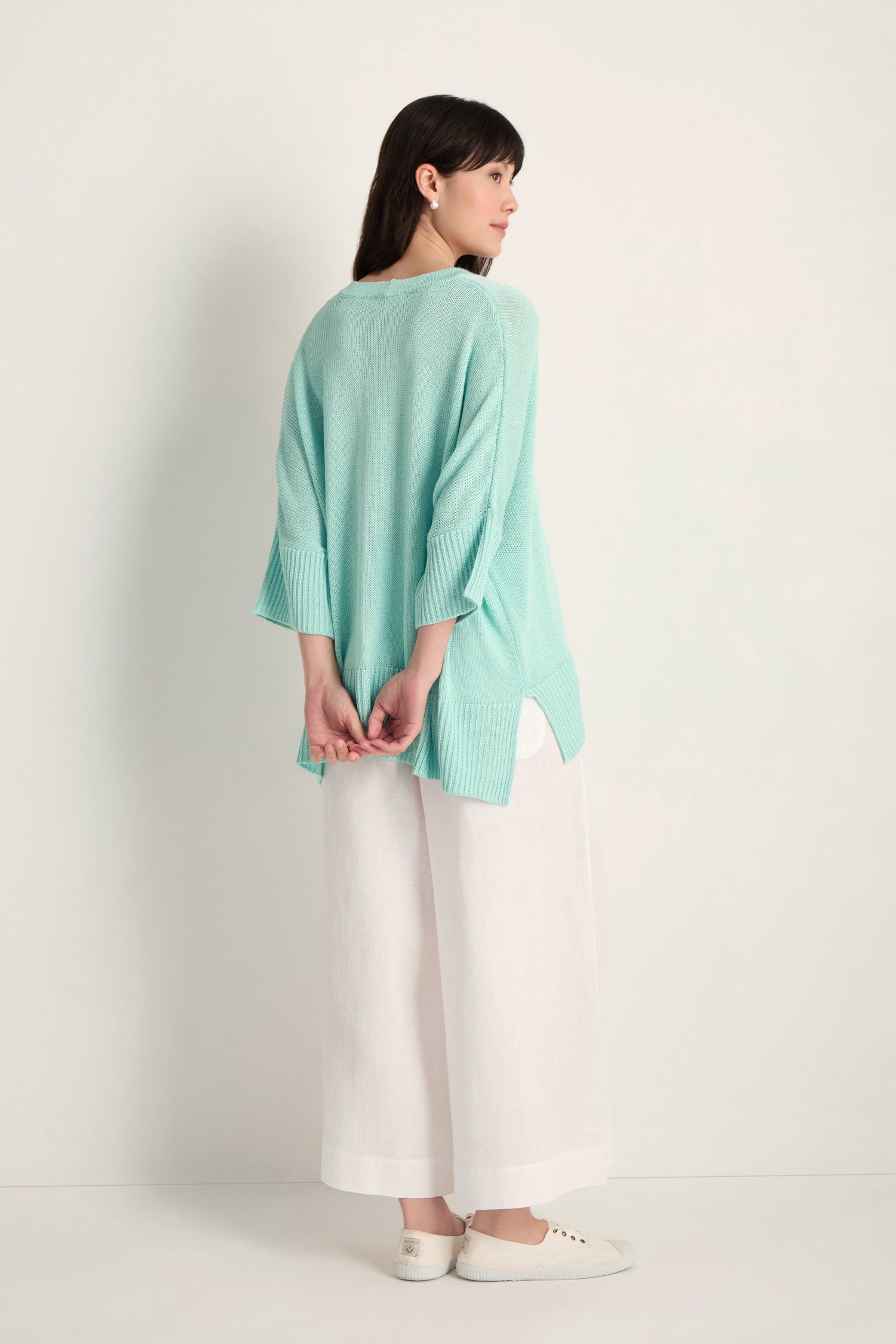 Facing away, a woman wears the Relaxed Aqua Cardigan, styled with wide-leg white pants and white sneakers against a plain light background.