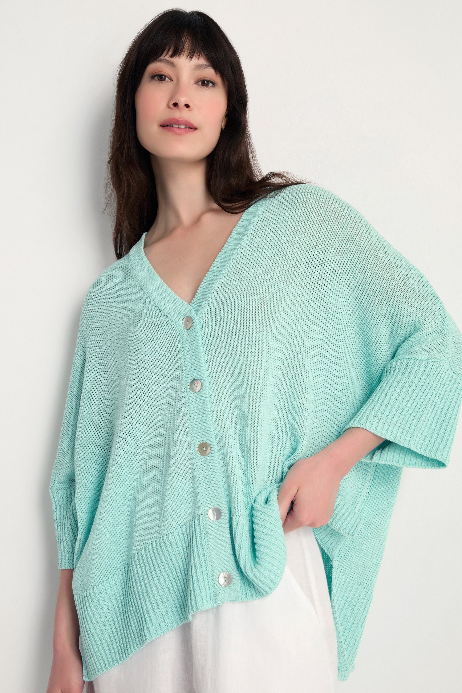 A woman wears the Relaxed Aqua Cardigan, a light blue open knit style with large buttons, layered over a white outfit against a plain background—ideal for lightweight layering.
