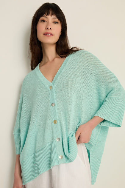 A woman wears the Relaxed Aqua Cardigan, a light blue open knit style with large buttons, layered over a white outfit against a plain background—ideal for lightweight layering.