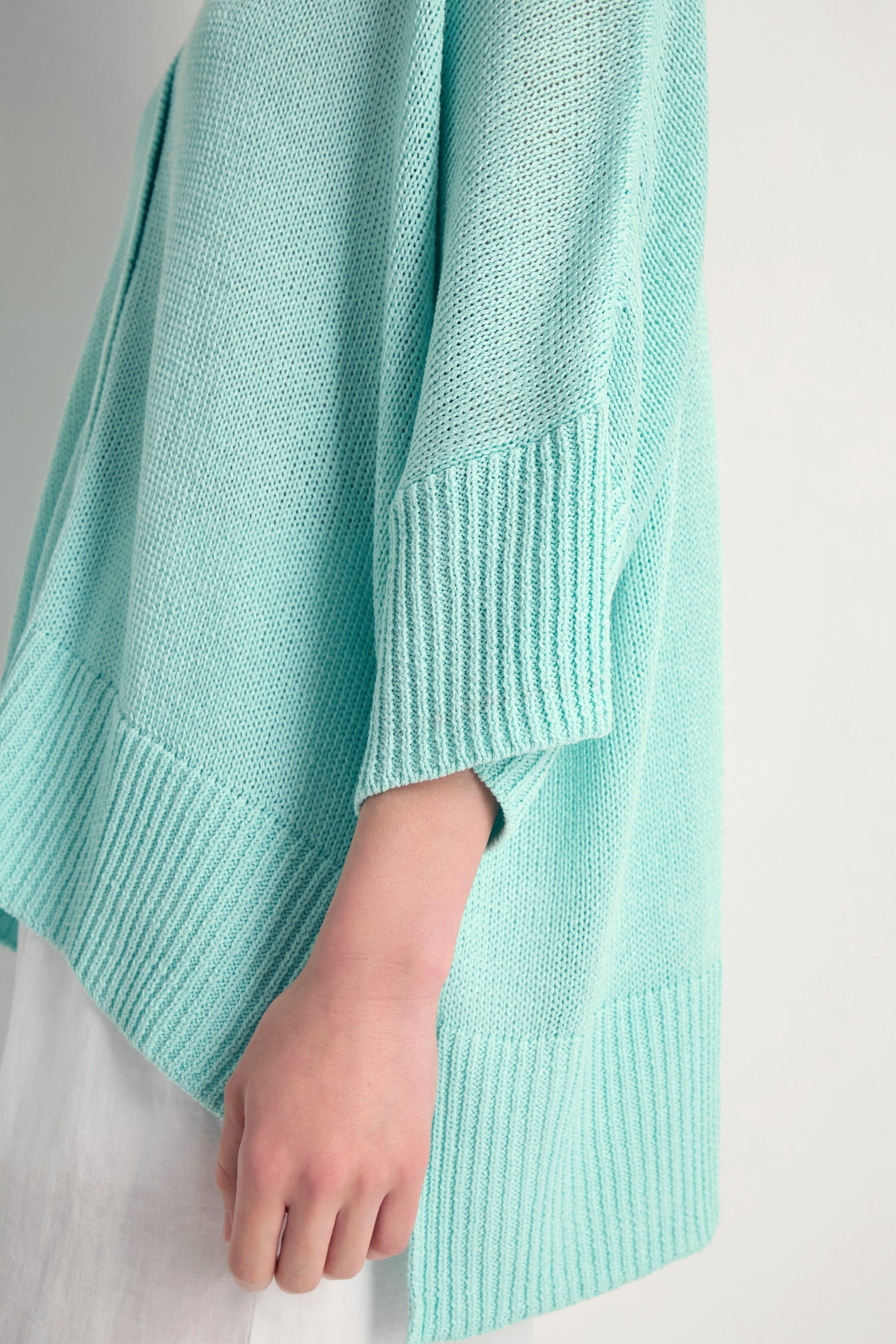 A person wearing the Relaxed Aqua Cardigan—a light blue open knit sweater with ribbed cuffs and hem—styled with white pants and shown from the side, perfect for lightweight layering.