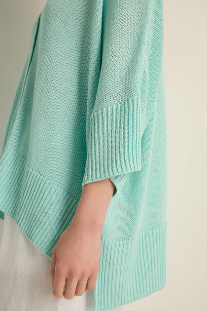 A person wearing the Relaxed Aqua Cardigan—a light blue open knit sweater with ribbed cuffs and hem—styled with white pants and shown from the side, perfect for lightweight layering.