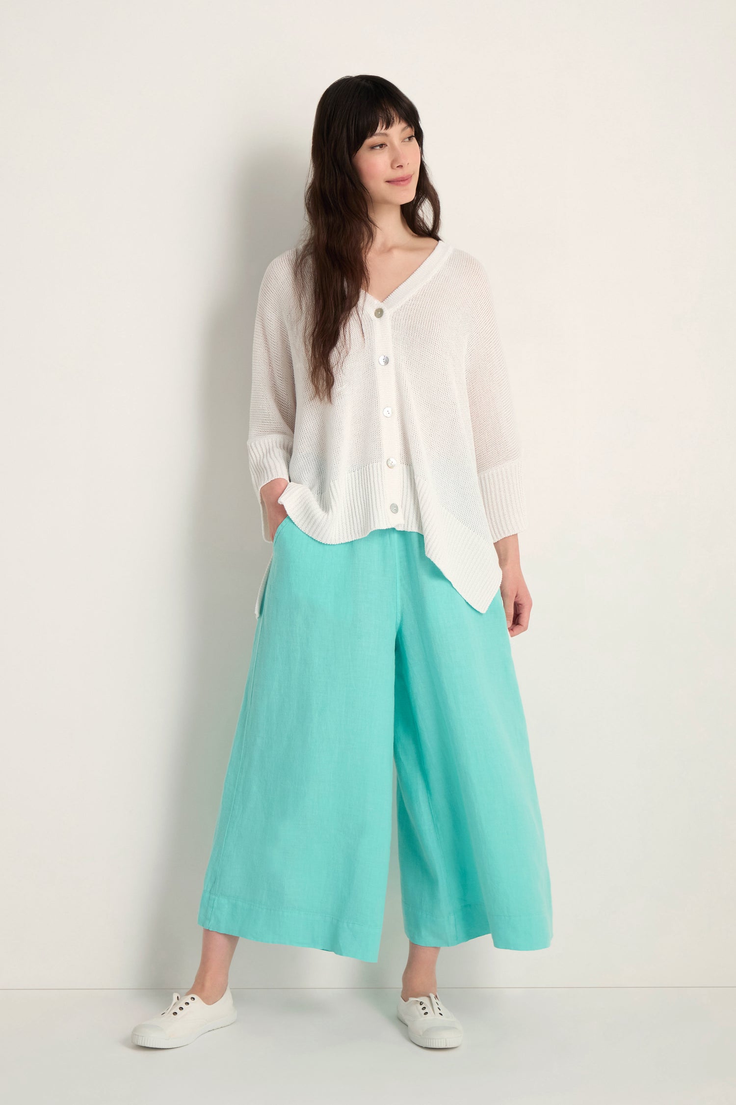 A woman stands against a plain background wearing the Relaxed White Cardigan, ideal for lightweight layering, styled with light blue wide-leg pants and white sneakers.