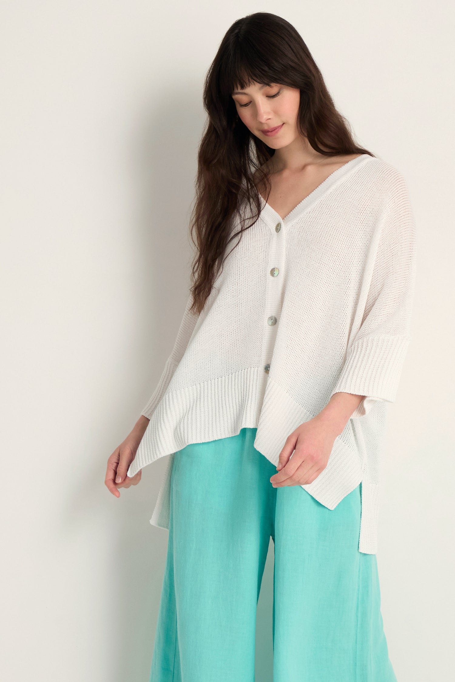 A woman wears the Relaxed White Cardigan with an asymmetric hem and wide light blue pants, standing against a plain light background—perfect for lightweight layering.