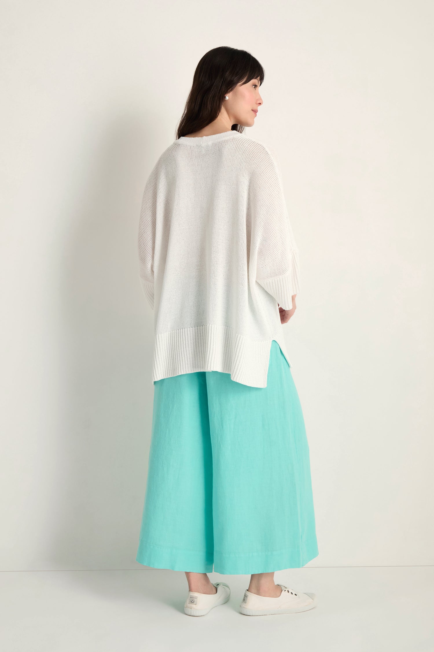 A woman stands against a plain background, facing away. She wears the Relaxed White Cardigan, turquoise wide-leg pants, and white slip-on shoes.