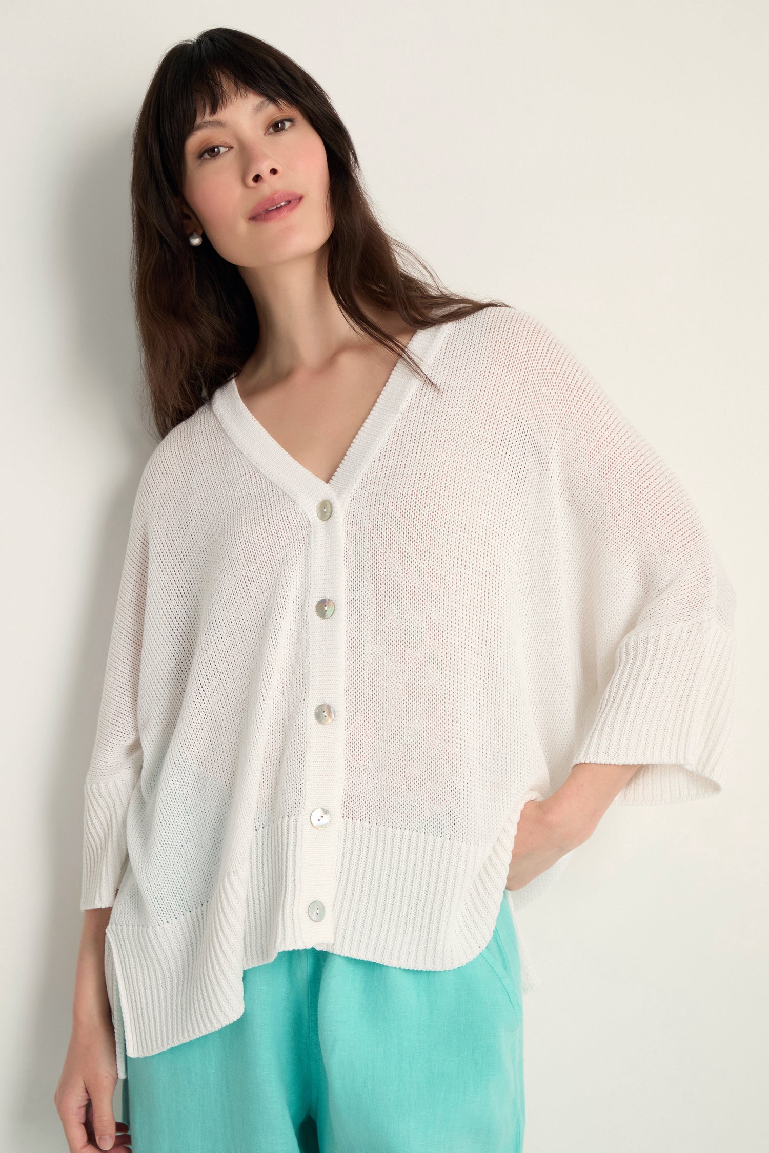 Woman wears the Relaxed White Cardigan—a loose, open-knit white button-up—paired with light blue pants against a plain background. Perfect lightweight layering piece.