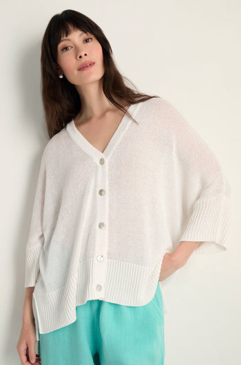 Woman wears the Relaxed White Cardigan—a loose, open-knit white button-up—paired with light blue pants against a plain background. Perfect lightweight layering piece.