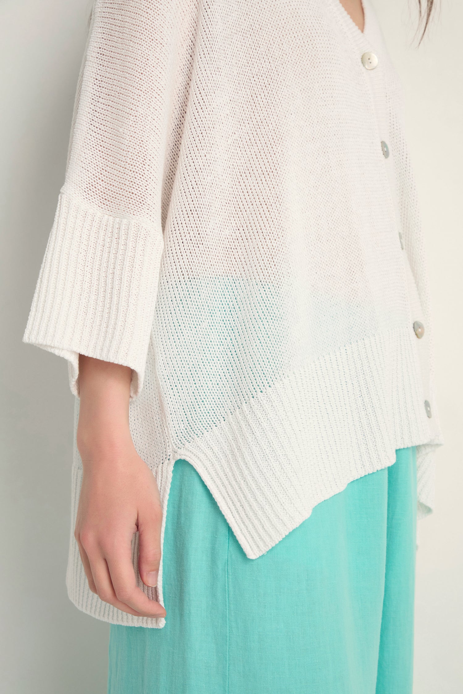 A person is shown from the shoulders down wearing the Relaxed White Cardigan—an open knit white sweater with large buttons—styled over a light turquoise skirt.
