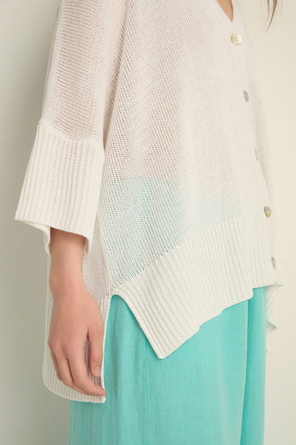 A person is shown from the shoulders down wearing the Relaxed White Cardigan—an open knit white sweater with large buttons—styled over a light turquoise skirt.