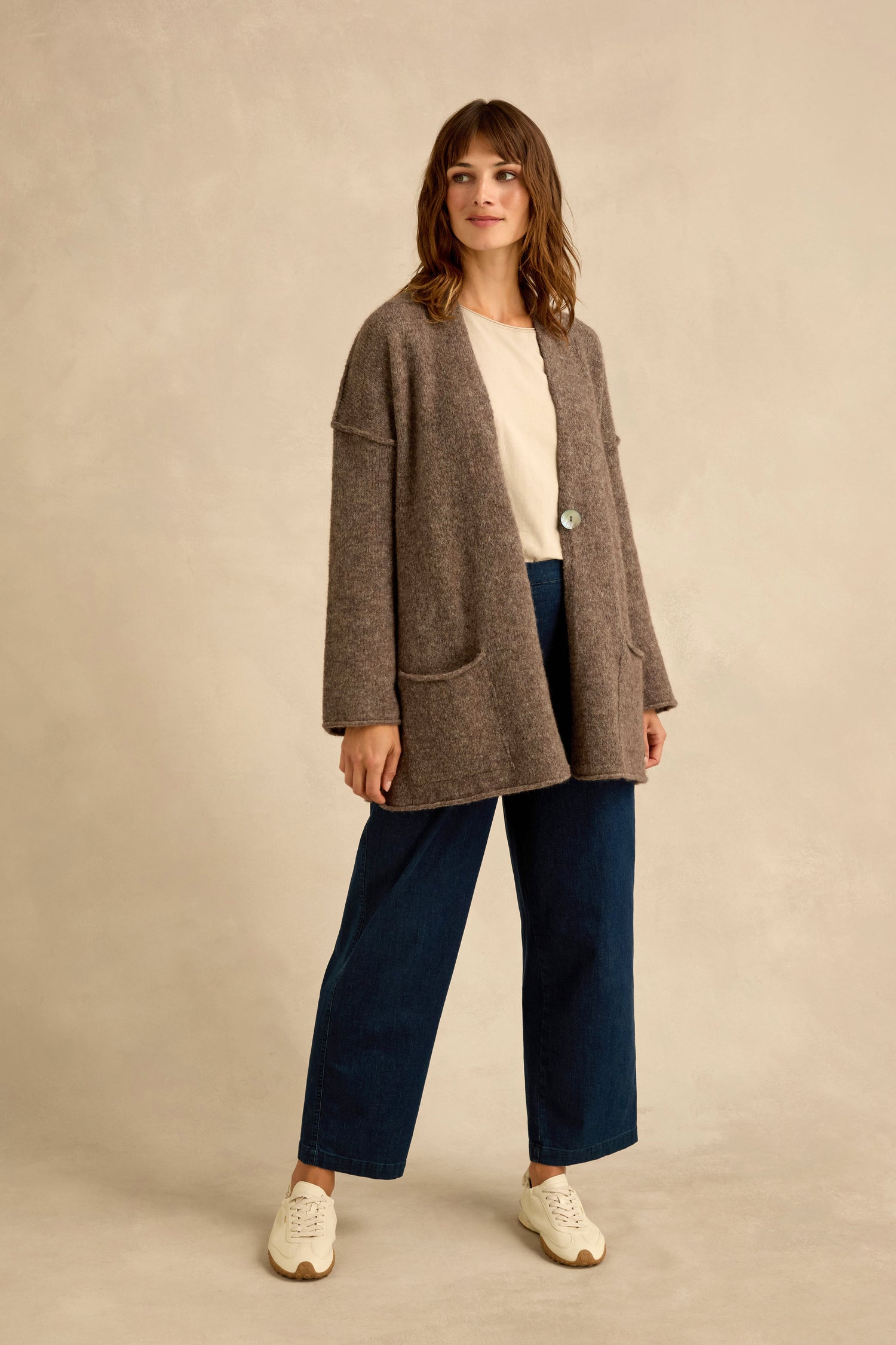 A woman stands against a beige background, wearing a beige top, wide-leg blue jeans, white sneakers, and the Bark Alpaca Blend Long Line Cardigan.