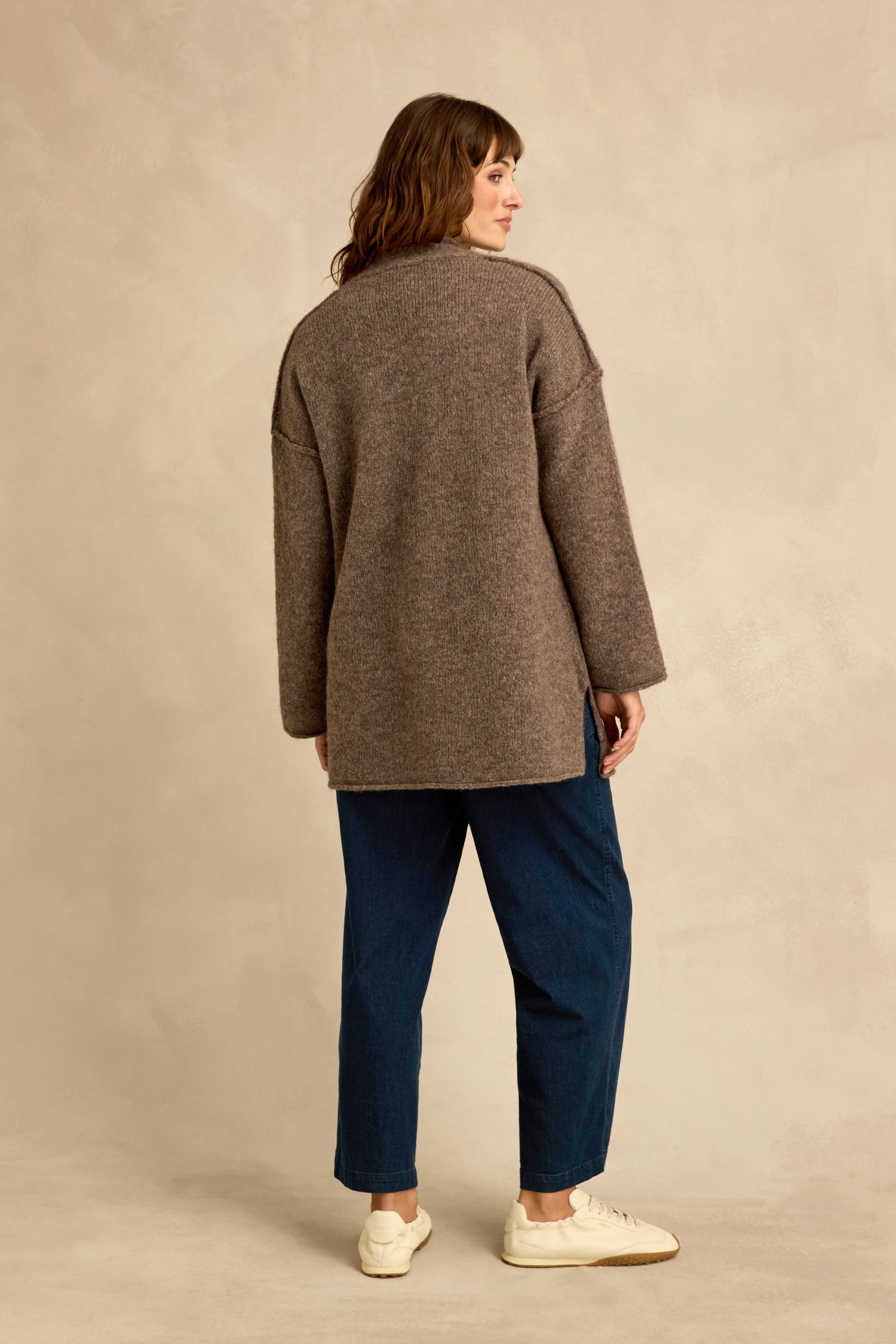 A woman stands facing away, wearing the Bark Alpaca Blend Long Line Cardigan with blue jeans and white sneakers against a neutral background.