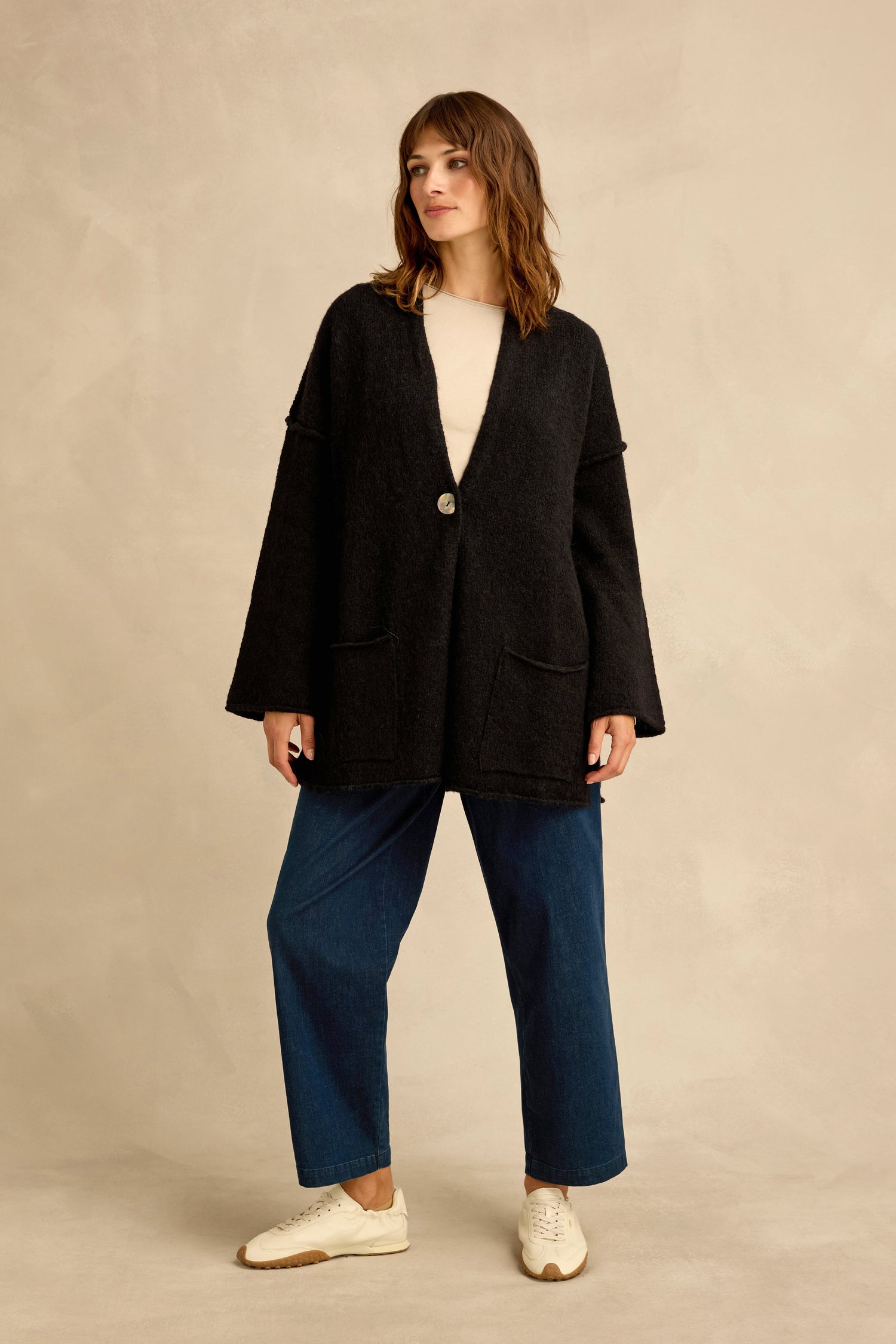 A person stands against a neutral background wearing the Black Alpaca Blend Long Line Cardigan, a cream top, blue wide-leg jeans, and white sneakers—a stylish take on modern women’s knitwear.