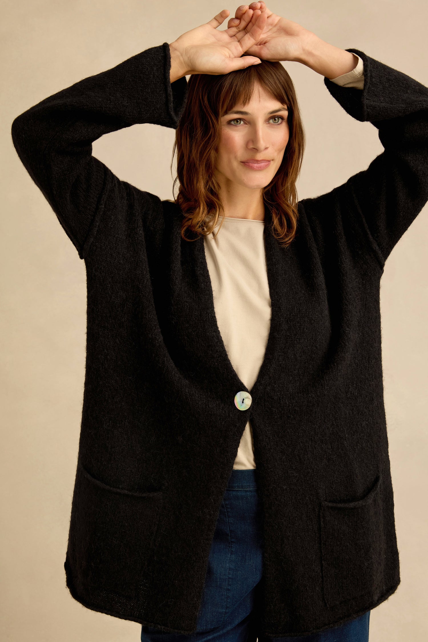 A woman stands with her arms raised, wearing the Black Alpaca Blend Long Line Cardigan over a beige top and blue pants, set against a plain neutral background.