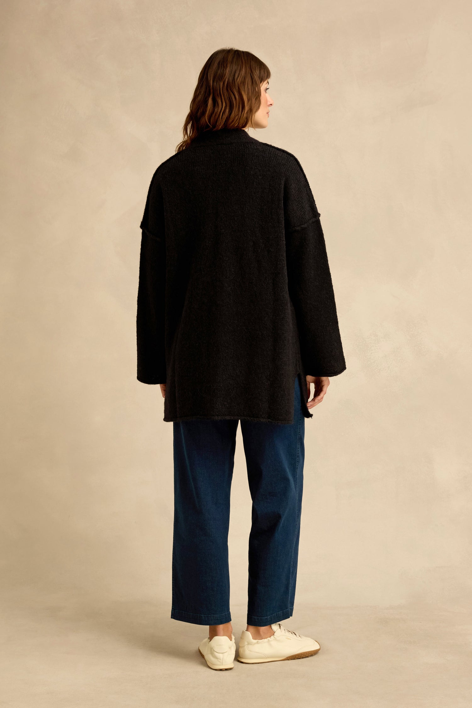 A person stands with their back to the camera, wearing the Black Alpaca Blend Long Line Cardigan over blue wide-leg pants and white sneakers against a neutral background.