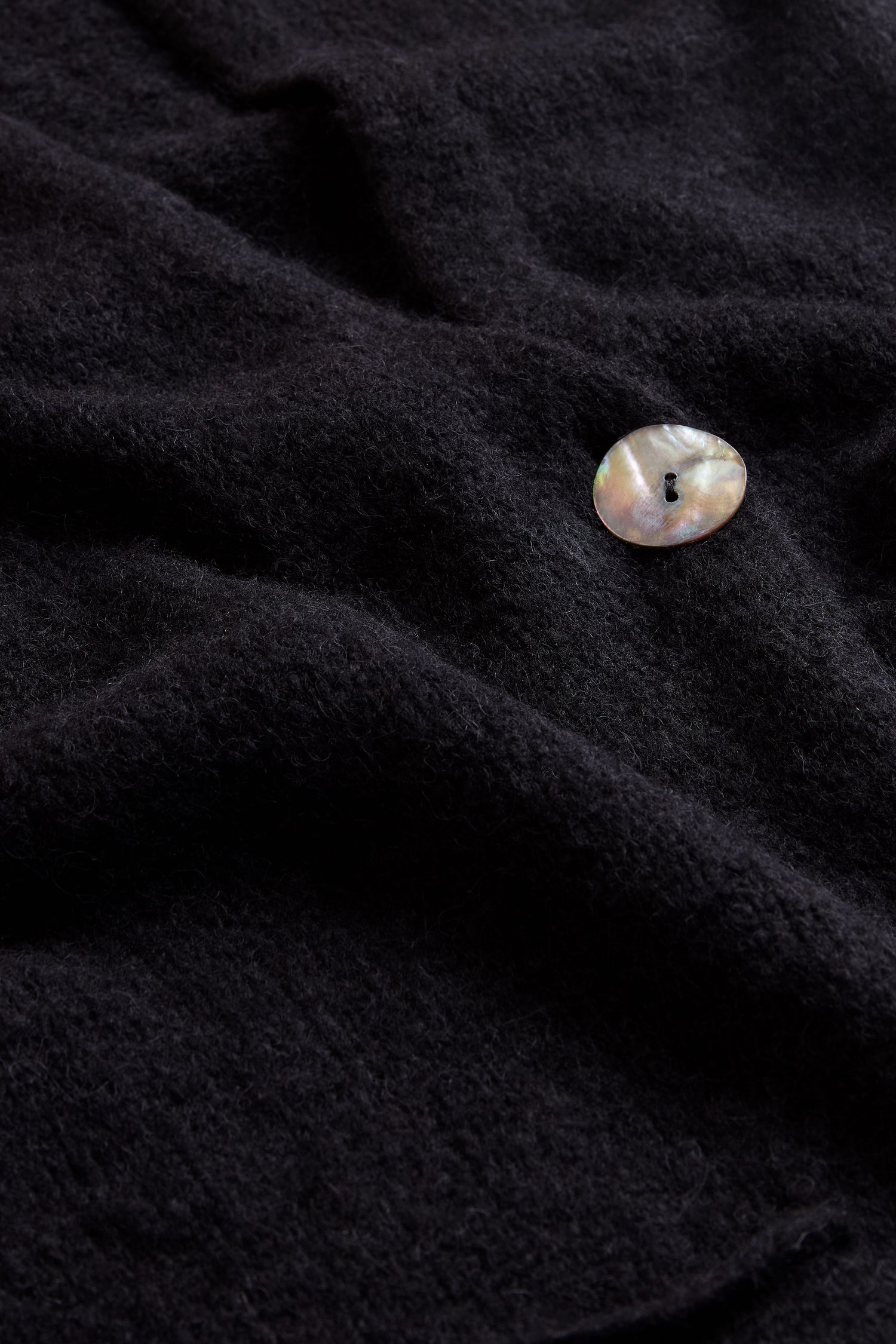 A single iridescent button rests on the softly textured, dark surface of the Black Alpaca Blend Long Line Cardigan, its visible folds and woolly finish evoking a luxurious touch.