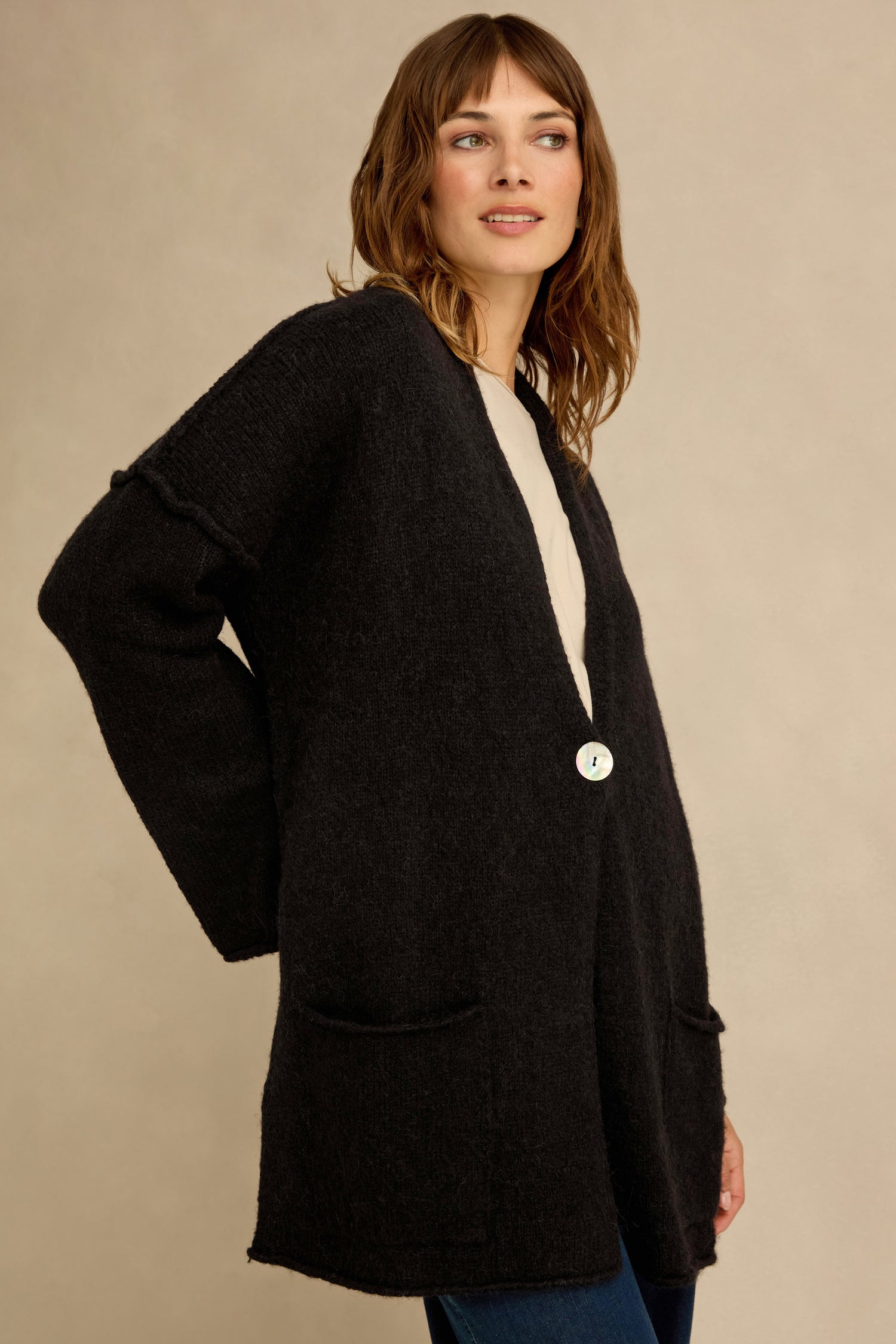 A woman is wearing the Black Alpaca Blend Long Line Cardigan, featuring front pockets and a single white button, standing against a beige background.