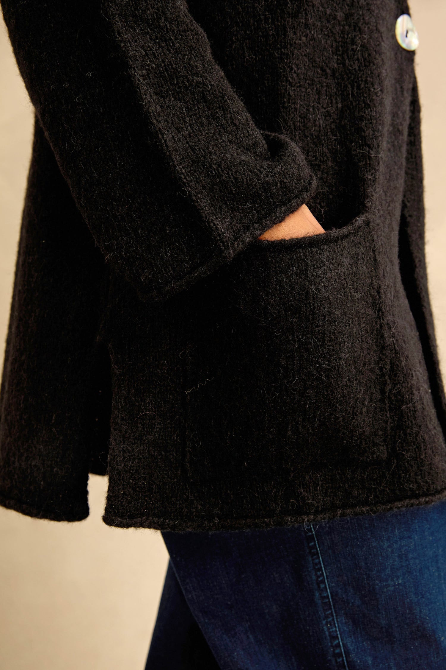 A person wearing the Black Alpaca Blend Long Line Cardigan, featuring a textured black finish and a hand in the pocket, paired with blue jeans and shown from the side.