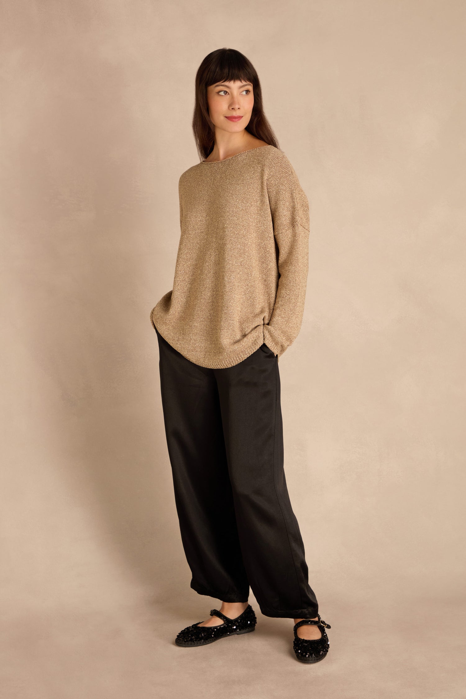 Woman stands against a beige background wearing the Champagne Boxy Sparkle Knit Jumper, black pants and shoes, hands in pockets, gazing to the side.