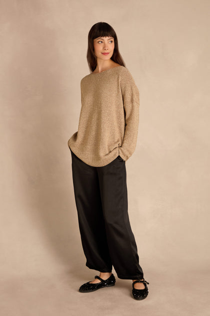 Woman stands against a beige background wearing the Champagne Boxy Sparkle Knit Jumper, black pants and shoes, hands in pockets, gazing to the side.