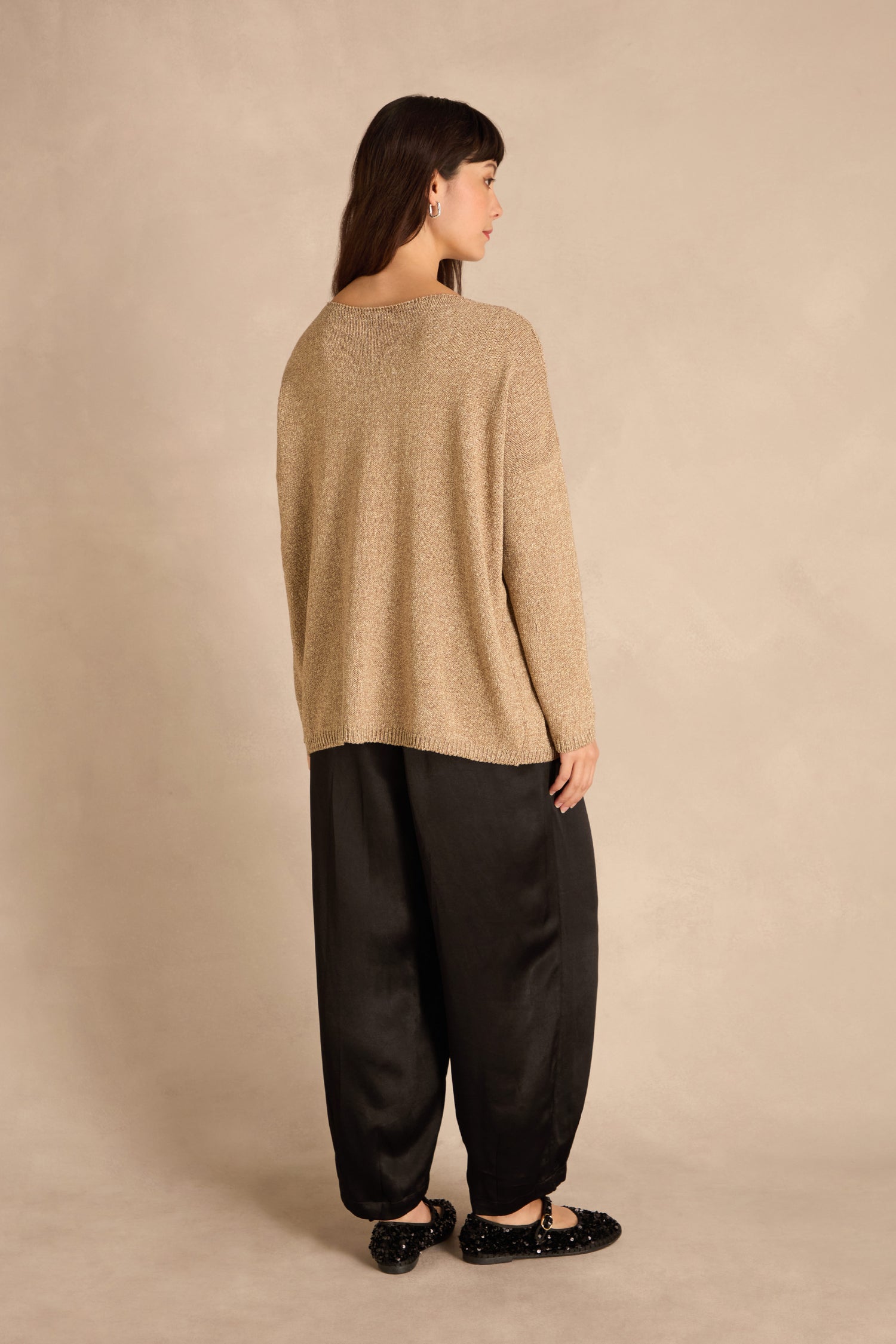 A woman faces away, wearing the Champagne Boxy Sparkle Knit Jumper, black wide-leg pants, and black shoes against a plain beige background.