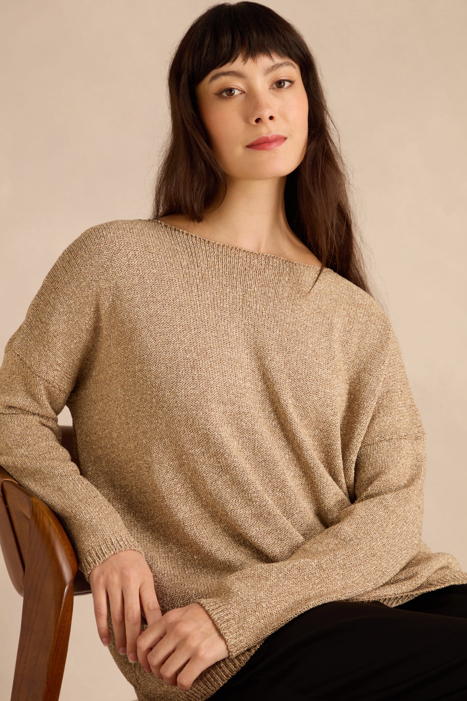 Woman with long dark hair sits on a wooden chair, wearing the Champagne Boxy Sparkle Knit Jumper and black pants, looking at the camera against a plain background.