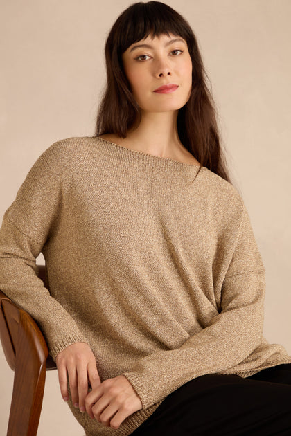 Woman with long dark hair sits on a wooden chair, wearing the Champagne Boxy Sparkle Knit Jumper and black pants, looking at the camera against a plain background.