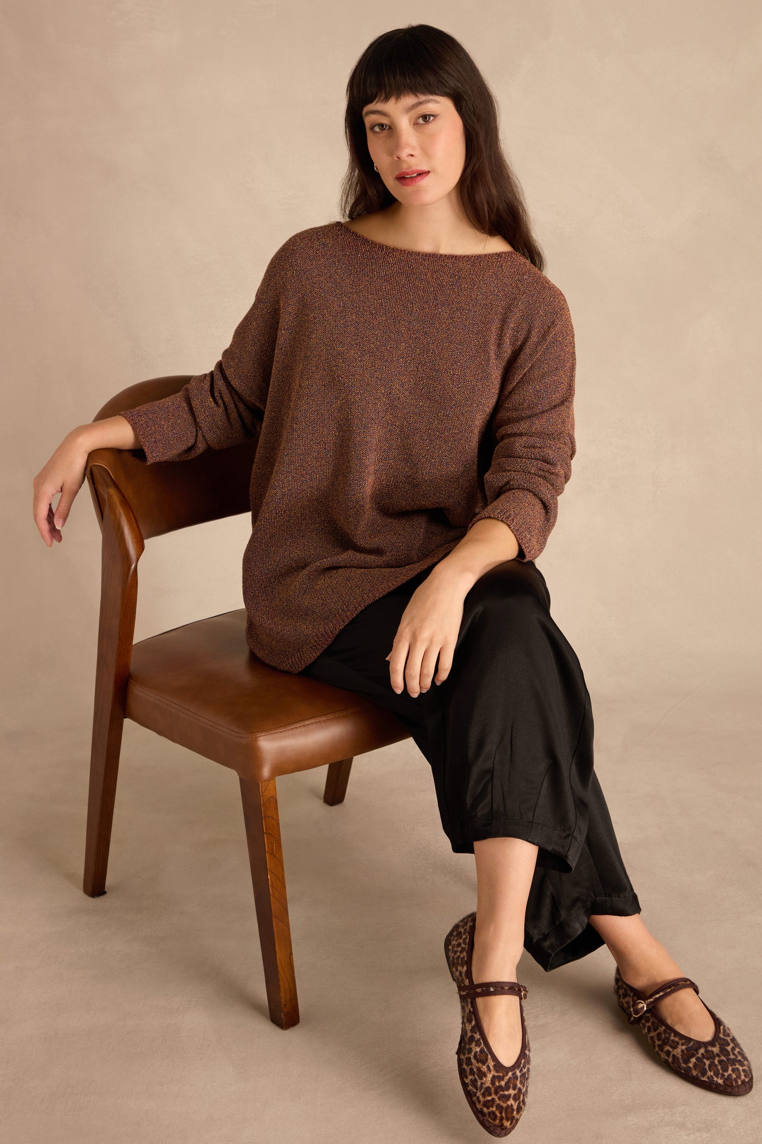 Woman sitting sideways on a brown chair in the Copper Boxy Sparkle Knit Jumper, black pants, and leopard print shoes, one arm resting on the chair back.
