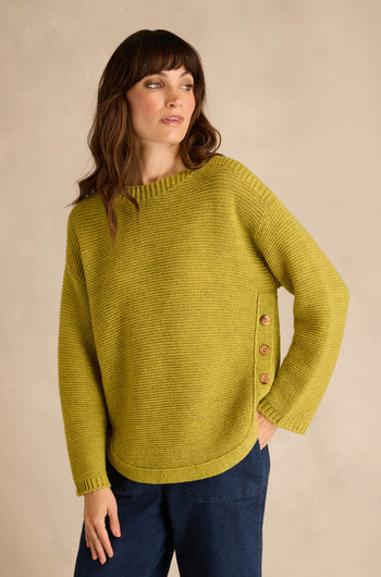 A woman wearing the Avocado Ribbed Wool Blend Button Jumper, a loose-fitting yellow-green top with side buttons, paired with dark blue pants and standing against a neutral background.
