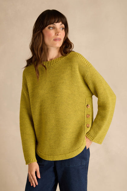 A woman wearing the Avocado Ribbed Wool Blend Button Jumper, a loose-fitting yellow-green top with side buttons, paired with dark blue pants and standing against a neutral background.