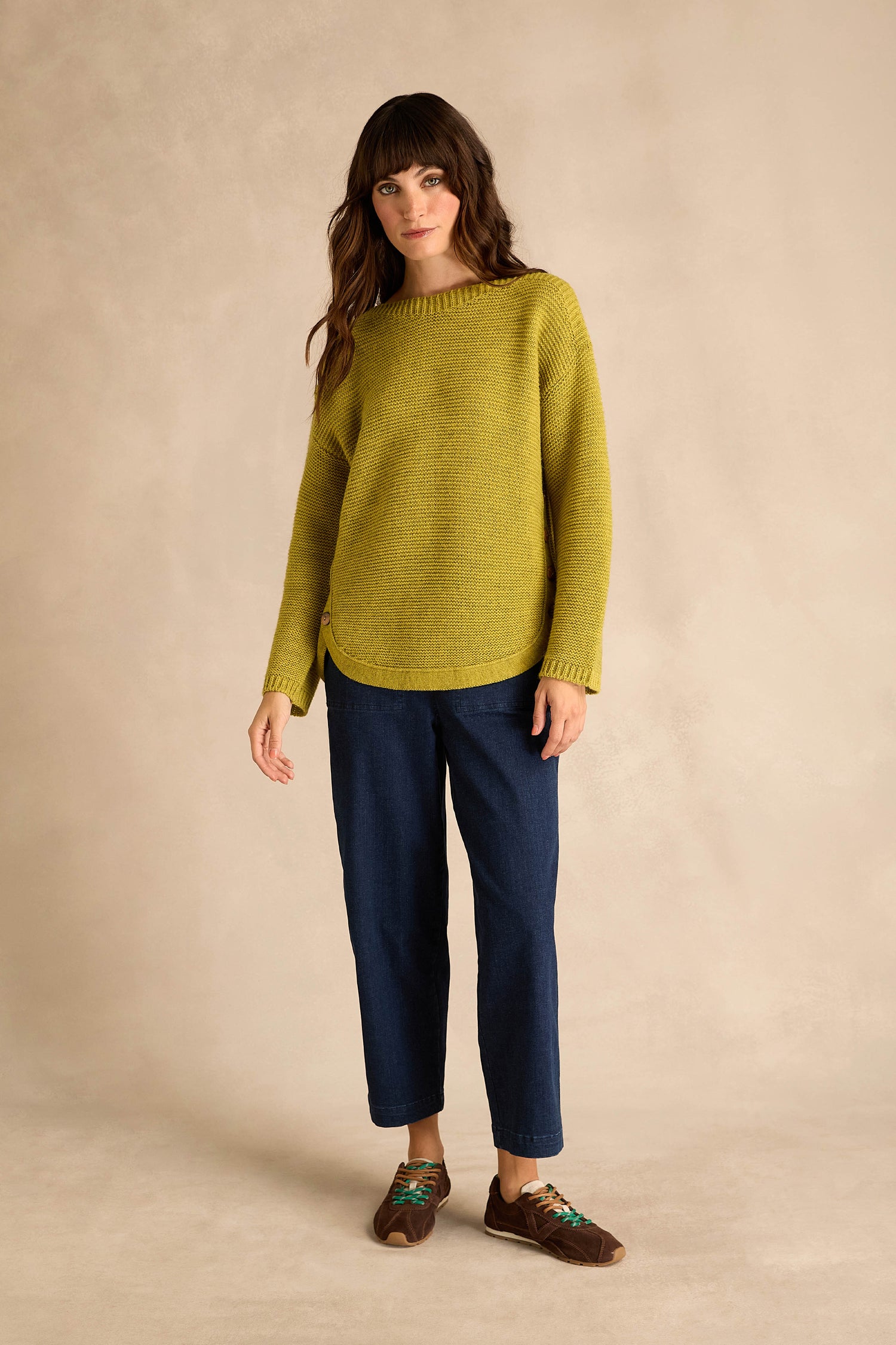 A woman stands against a beige backdrop, wearing the Avocado Ribbed Wool Blend Button Jumper, paired with dark blue pants and brown shoes.