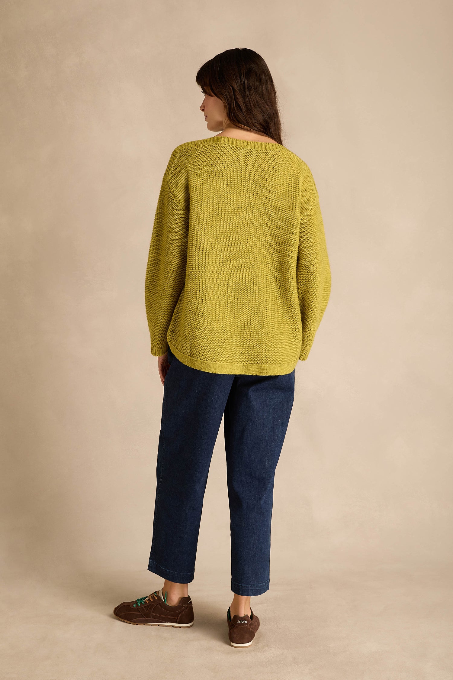 A person stands with their back to the camera, wearing the Avocado Ribbed Wool Blend Button Jumper—a loose yellow alpaca yarn sweater—paired with blue jeans and brown shoes, against a neutral beige background.