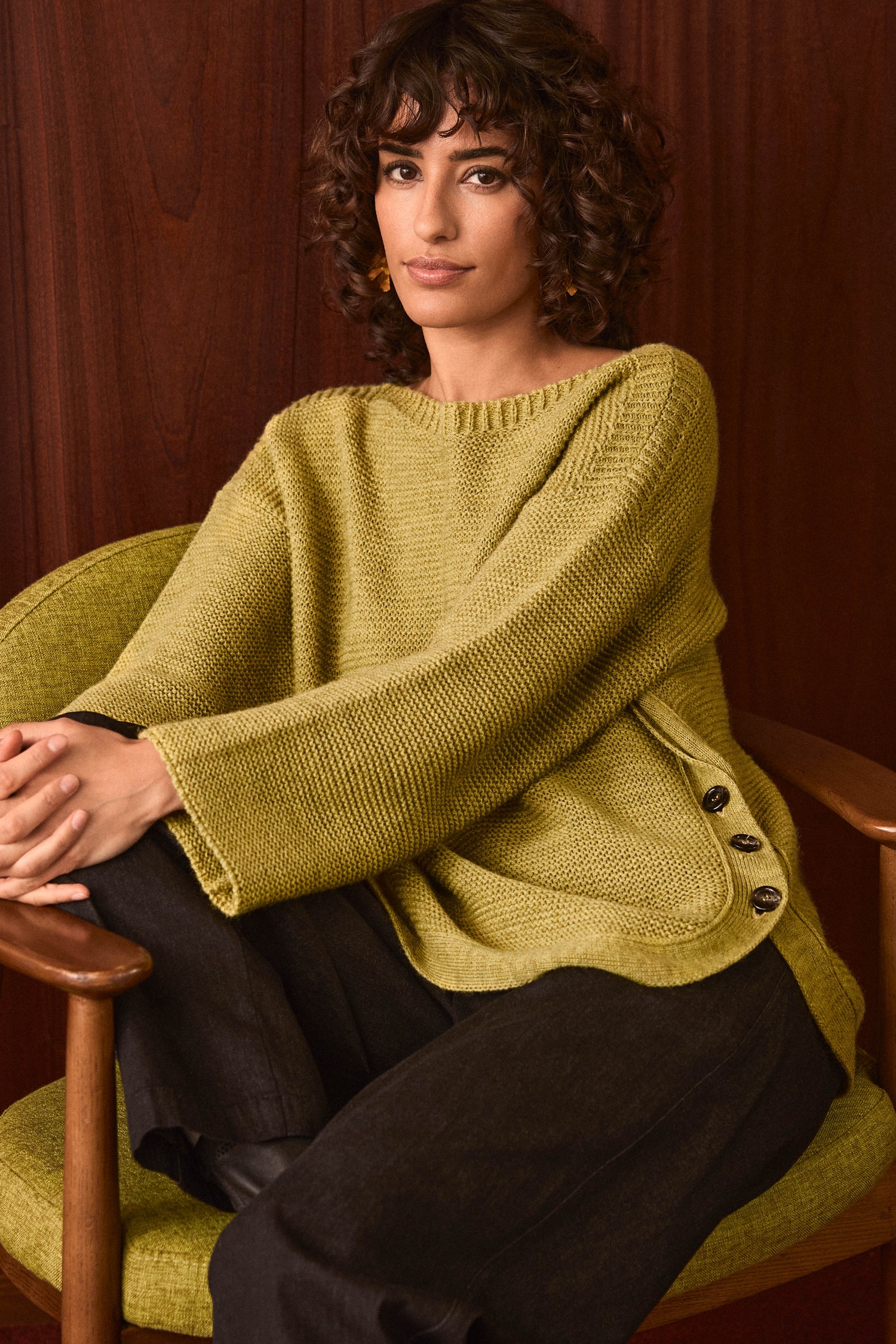 A woman with curly hair sits in a green chair against a wooden background, wearing the Avocado Ribbed Wool Blend Button Jumper—a cozy, loose olive knit with side buttons—paired with dark pants.