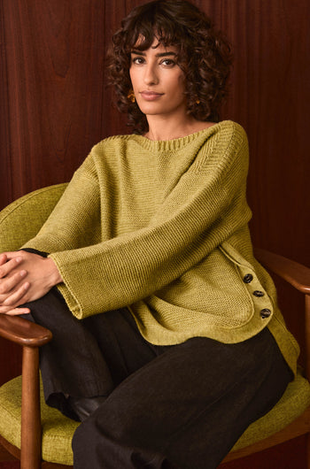 A woman with curly hair sits in a green chair against a wooden background, wearing the Avocado Ribbed Wool Blend Button Jumper—a cozy, loose olive knit with side buttons—paired with dark pants.