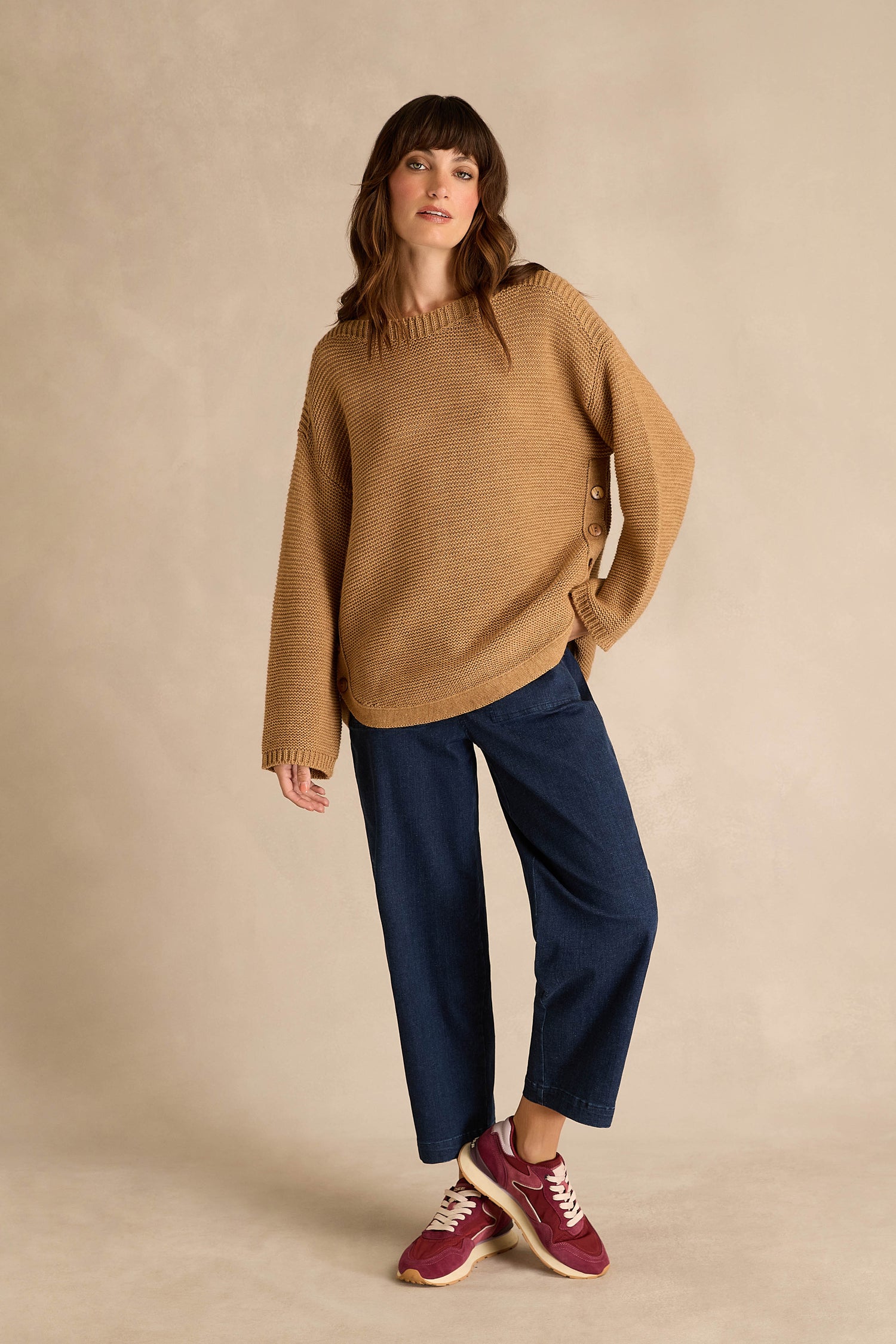 A woman wearing a Camel Ribbed Wool Blend Button Jumper, blue wide-leg pants, and red sneakers stands against a plain beige background.