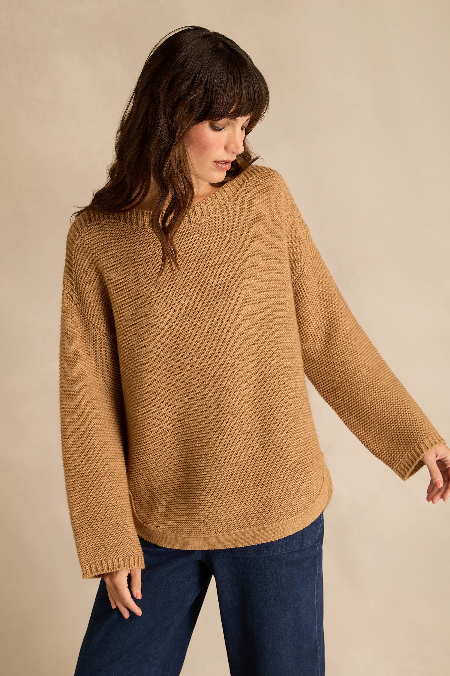 A person wearing the Camel Ribbed Wool Blend Button Jumper stands against a beige background, looking down and to the side. The ribbed wool blend jumper pairs effortlessly with dark blue pants for an understated, classic look.